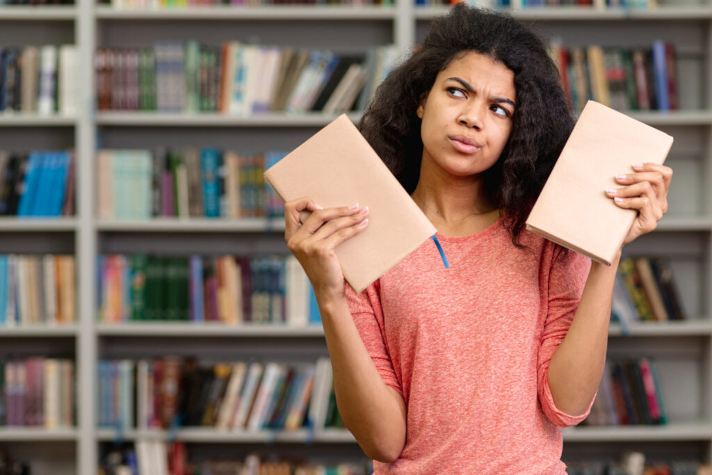 Mulher jovem de cabelos pretos e camiseta rosa segura um livro em cada mão com uma expressão de dúvida